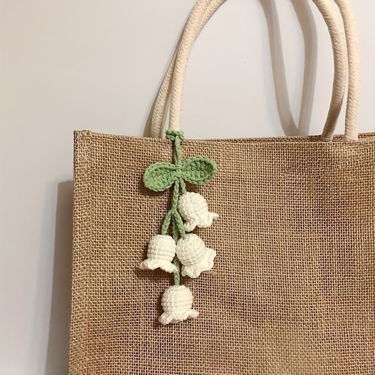 A jute tote bag with white rope handles, decorated with a handmade crochet charm featuring small white bell-shaped flowers and green leaves tied in a bow.