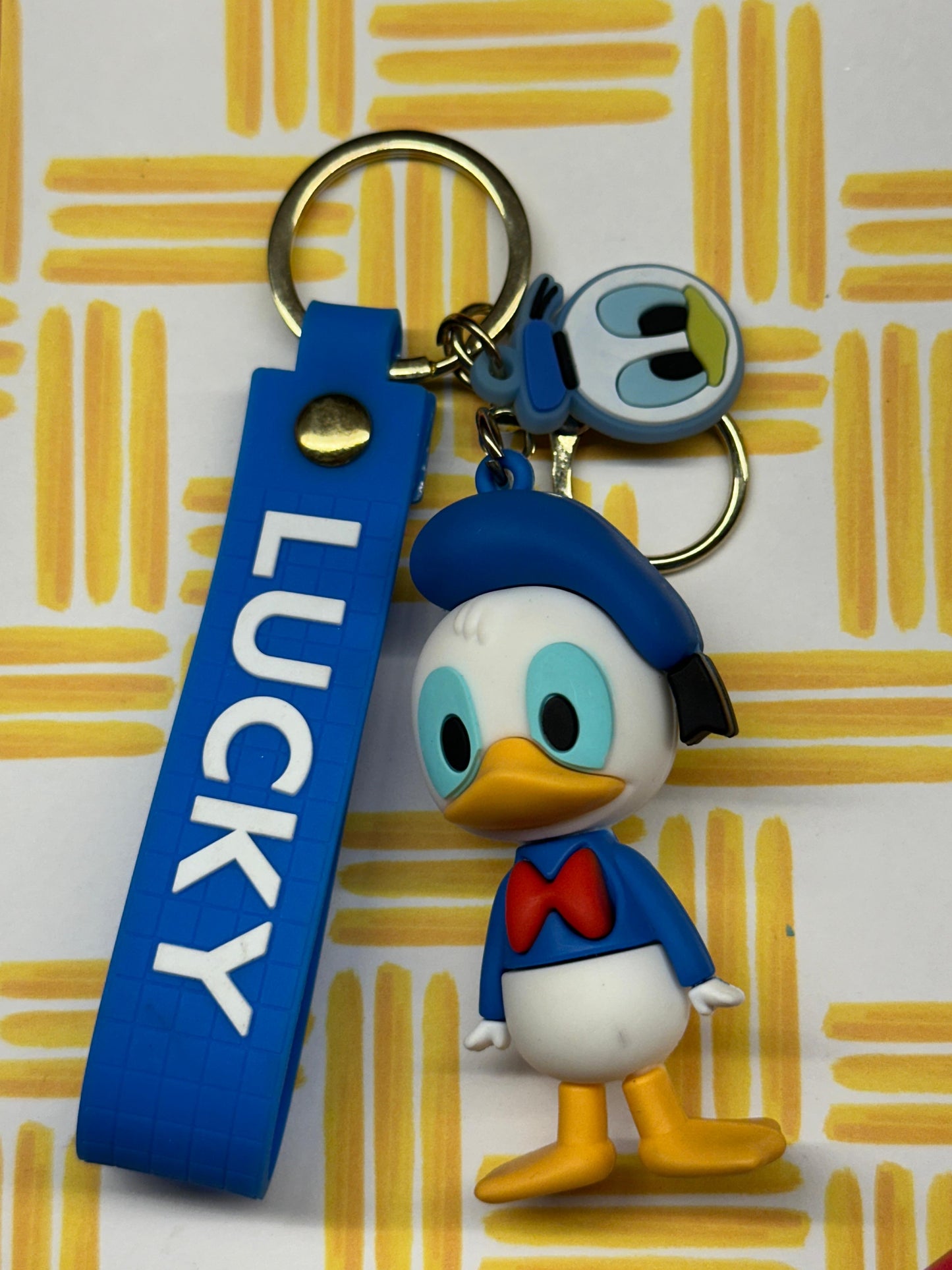 Donald Duck keychain with a blue “LUCKY” strap and a small face charm on a yellow patterned background.