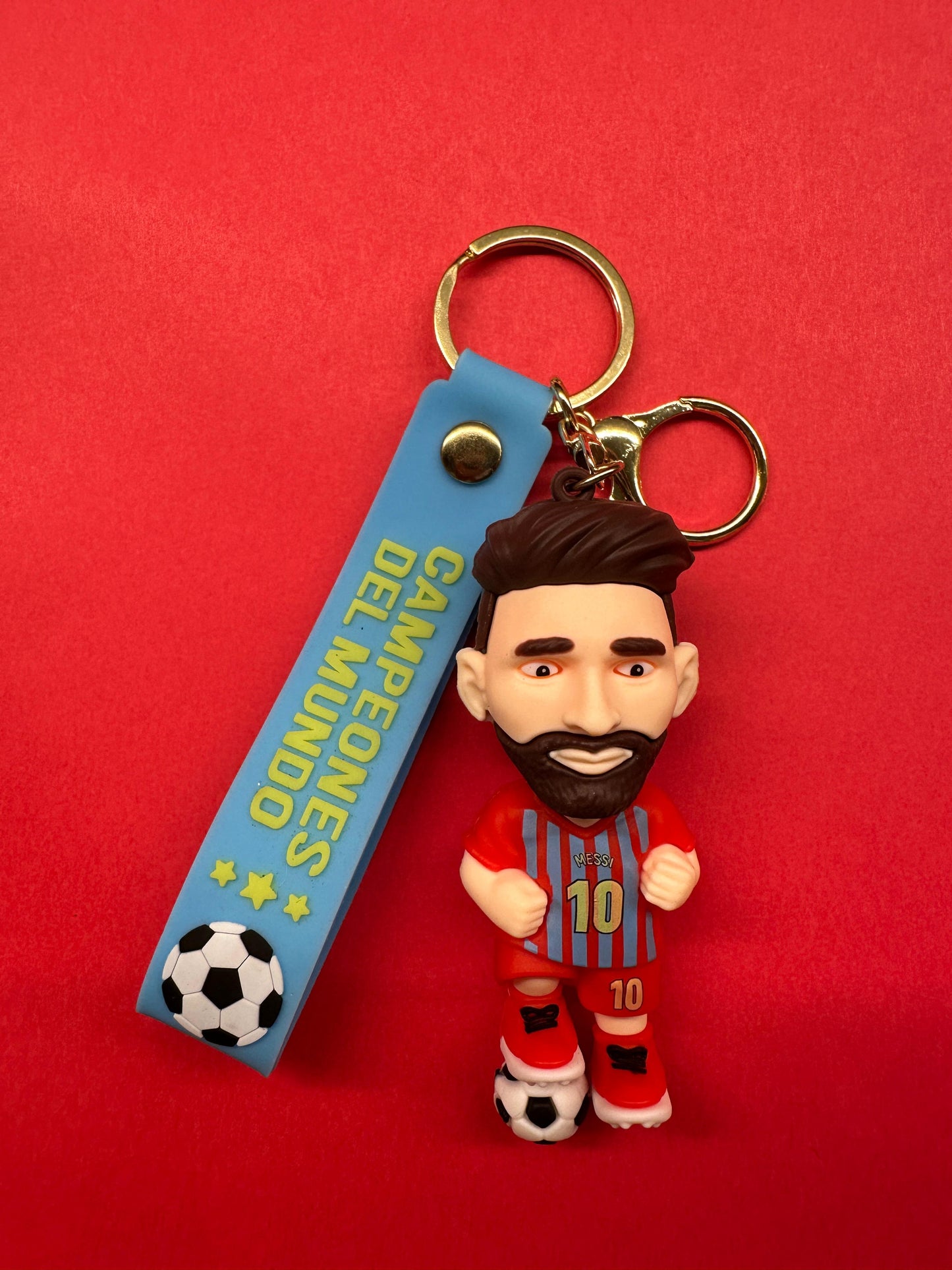 Messi-themed keychain with a cartoon figure in a red and blue striped jersey numbered 10, attached to a blue strap that says “CAMPEONES DEL MUNDO” with stars and a football icon, on a red background.