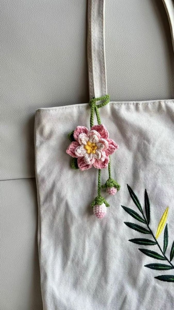Crocheted pink and yellow flower keychain dangling from the strap of a canvas tote bag, featuring a main flower and two smaller buds on a green stem.