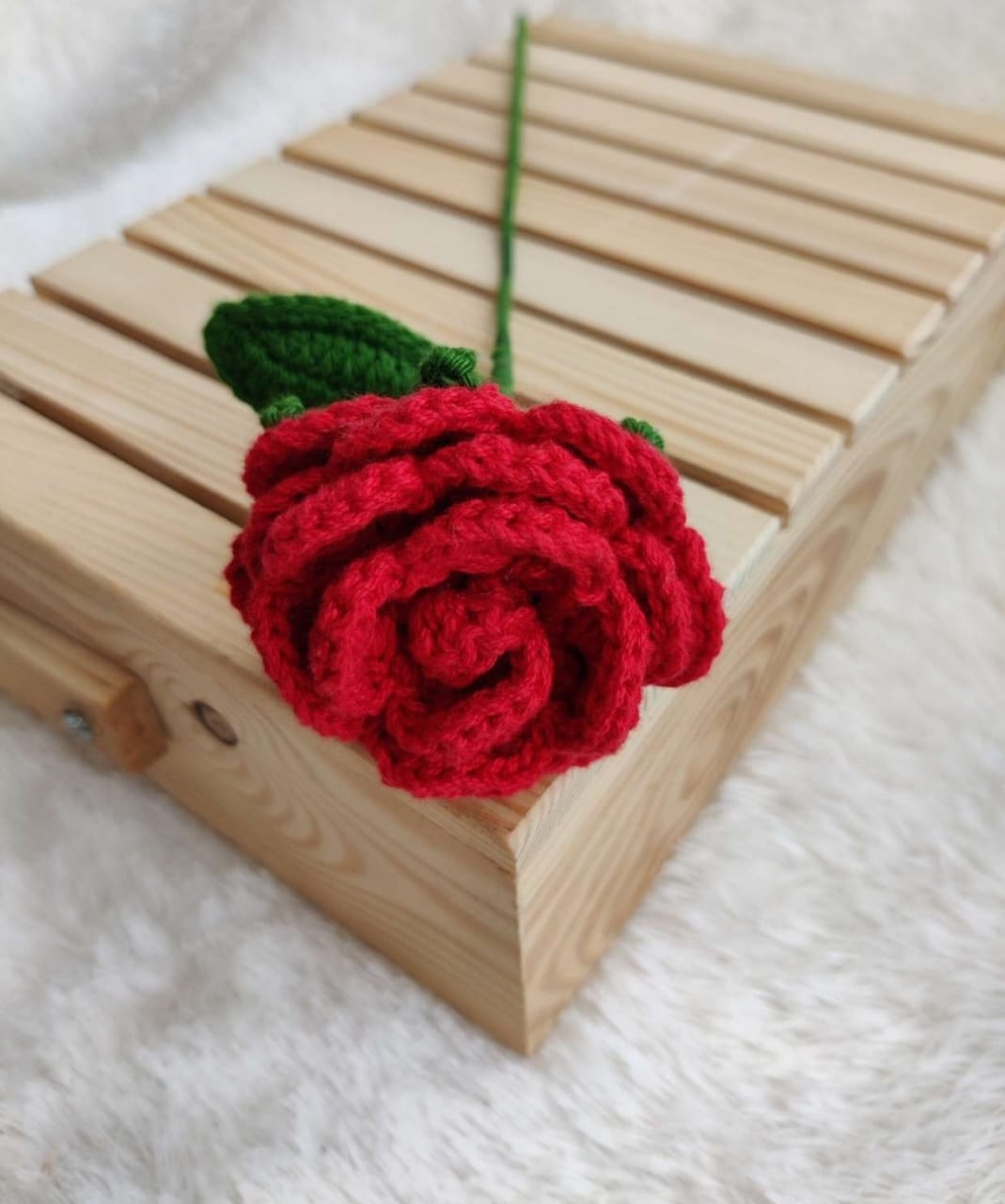 A handmade red crochet rose with a green stem and leaf, placed on a light wooden box against a soft white textured background.