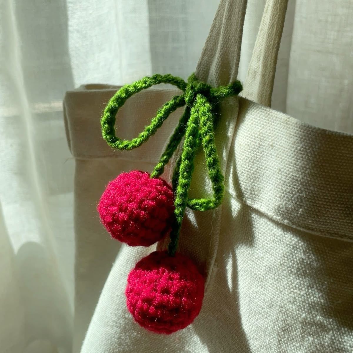 A cream-colored canvas tote bag decorated with a handmade crochet cherry charm featuring two red cherries and green stems tied in a bow.