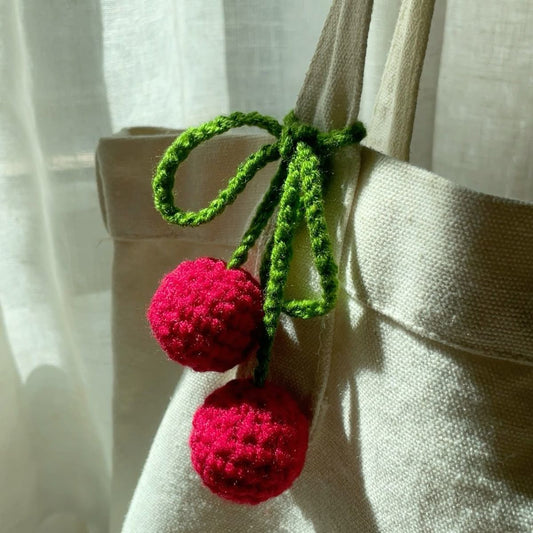 A cream-colored canvas tote bag decorated with a handmade crochet cherry charm featuring two red cherries and green stems tied in a bow.