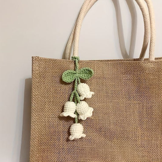 A jute tote bag with white rope handles, decorated with a handmade crochet charm featuring small white bell-shaped flowers and green leaves tied in a bow.