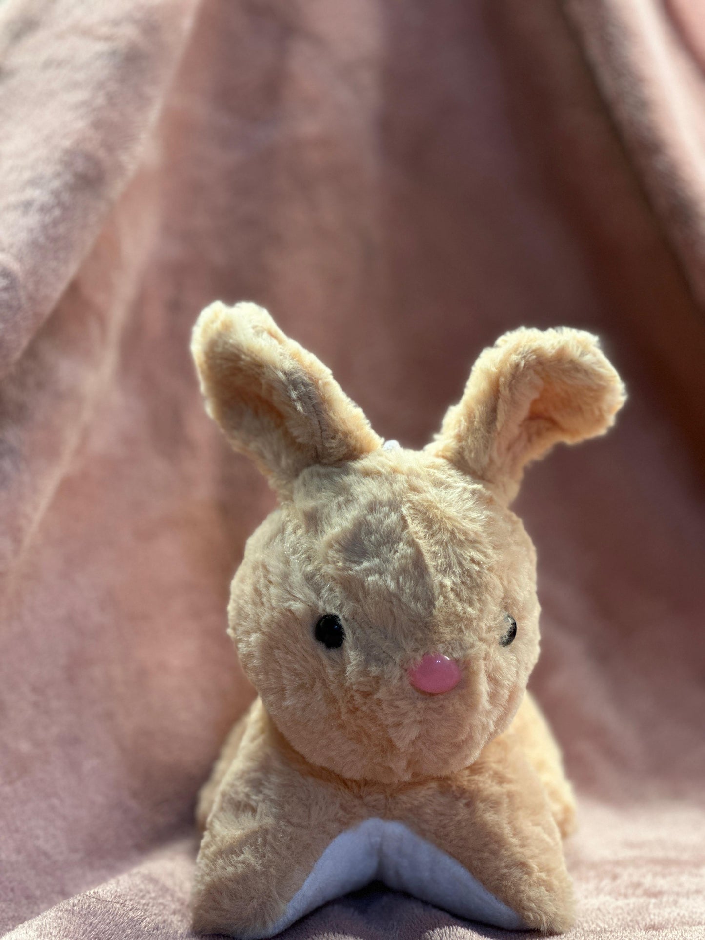  A cute beige plush bunny with black button eyes and a small pink nose, sitting on a soft pink fabric background. One of its ears is slightly bent, giving it an adorable and playful look.