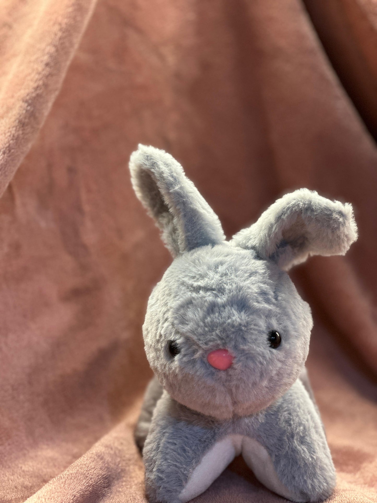  A cute grey plush bunny with black button eyes and a small pink nose, sitting on a soft pink fabric background. One of its ears is slightly bent, giving it an adorable and playful look.