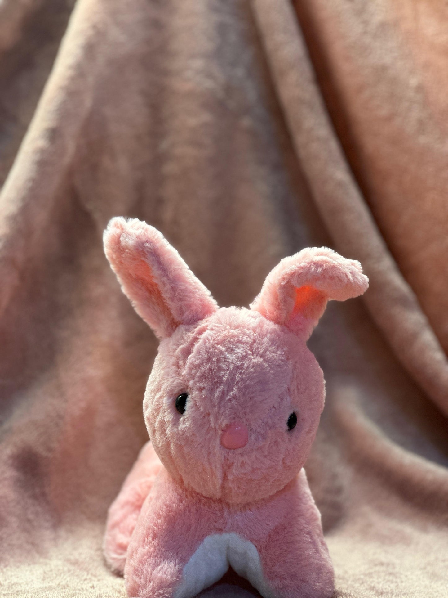  A cute pink plush bunny with black button eyes and a small pink nose, sitting on a soft pink fabric background. One of its ears is slightly bent, giving it an adorable and playful look.
