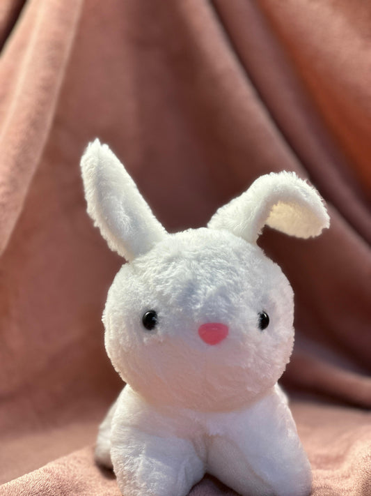  A cute white plush bunny with black button eyes and a small pink nose, sitting on a soft pink fabric background. One of its ears is slightly bent, giving it an adorable and playful look.