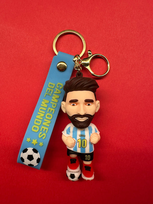 Messi-themed keychain showing a cartoon figure in Argentina’s blue and white jersey with number 10, attached to a blue strap reading “CAMPEONES DEL MUNDO” with stars and a football icon, on a red background.