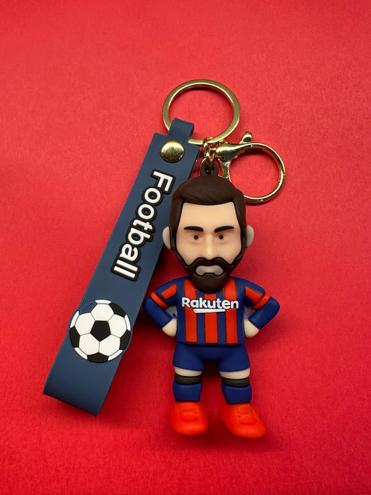 Miniature keychain of Messi in a Barcelona FC jersey and red boots, attached to a gold ring and a dark blue rubber strap labeled "Football" with a soccer ball graphic, all against a red background.